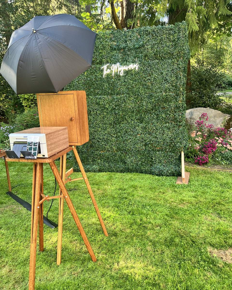 TBD's hand-built walnut photo booth on its wooden tripod, with studio flash umbrella and printer, set up on a lawn facing a greenery wall at an outdoor wedding