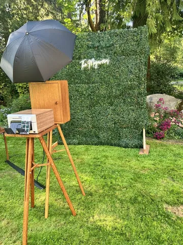 TBD's walnut photo booth set up on a lawn in front of a greenery wall at an outdoor wedding